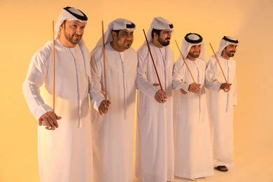 إيقاعات متناغمة وحركات جماعية تعكس روح الوحدة والاعتزاز بالهوية الوطنية،مجموعة من الرجال العرب الخليجيين الإماراتيين يرتدون الكندورة والغترة البيضاء يؤدون رقصة العيالة الشعبية بعصا الخيزران، أجواء تراثية تعبّر عن الفخر بالموروث الثقافي الإماراتي، الحفاظ على التراث الشعبي في المناسبات الوطنية والاجتماعية.
