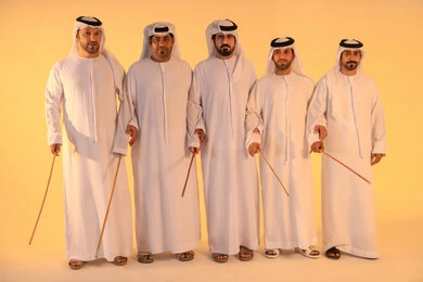 Harmonious rhythms and collective movements reflect the spirit of unity and pride in national identity. A group of Emirati Gulf Arab men wearing the kandura and white ghutrah perform the traditional Ayala dance with bamboo sticks. The atmosphere is cultural, expressing pride in the Emirati cultural heritage and preserving the folk heritage during national and social occasions.