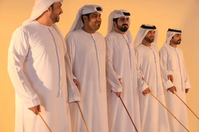 أجواء تراثية تعبّر عن الفخر بالموروث الثقافي الإماراتي،إيقاعات متناغمة وحركات جماعية تعكس روح الوحدة والاعتزاز بالهوية الوطنية،مجموعة من الرجال العرب الخليجيين الإماراتيين يرتدون الكندورة والغترة البيضاء يؤدون رقصة العيالة الشعبية بعصا الخيزران،  الحفاظ على التراث الشعبي في المناسبات الوطنية والاجتماعية.