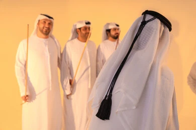 A group of Arab Gulf Emirati men wearing kandura and white ghutras perform the traditional Ayala dance with bamboo sticks. The atmosphere is cultural, expressing pride in the Emirati cultural heritage, with harmonious rhythms and collective movements reflecting the spirit of unity and pride in national identity, preserving the folk heritage during national and social occasions.