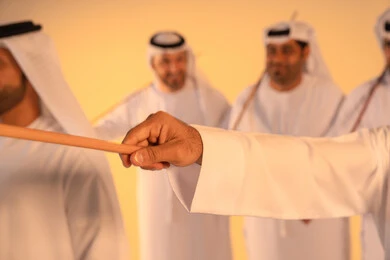 Embodiment of pride in Emirati customs and traditions, a close-up portrait of the hand of an Emirati Gulf Arab man wearing the kandura and ghutrah performing the Ayala dance with a bamboo stick, a joyful festive atmosphere, courage, generosity, and national belonging, preserving the popular heritage and cultural identity in national and social occasions.