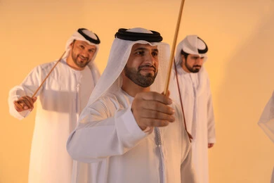 Embodiment of pride in Emirati customs and traditions, an Arab Gulf Emirati man wearing the kandura and ghutrah performs the Ayala dance with a bamboo stick, in a joyful festive atmosphere, showcasing courage, generosity, and national belonging, preserving the popular heritage and cultural identity during national and social occasions.
