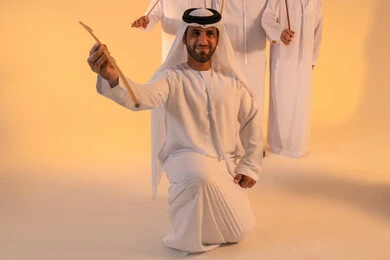 Embodiment of pride in Emirati customs and traditions, an Arab Gulf Emirati man wearing the kandura and ghutrah performs the Ayala dance with a bamboo stick, in a joyful festive atmosphere, showcasing courage, generosity, and national belonging, preserving the popular heritage and cultural identity during national and social occasions.