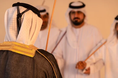 الحفاظ على العادات والتقاليد الشعبية في المناسبات الوطنية، مظاهر الفرح والاحتفاء بالتراث الإماراتي الأصيل، بورتريه لمجموعة من الرجال العرب الخليجيين الإماراتيين يرتدون الكندورة والغترة يؤدون رقصة العيالة التقليدية بالعصي الخشبية، تجسيد للهوية الوطنية والانتماء، أجواء احتفالية مفعمة بالفخر والاعتزاز.