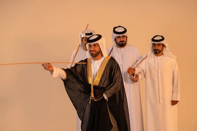 تجسيد للهوية الوطنية والانتماء، الحفاظ على العادات والتقاليد الشعبية في المناسبات الوطنية، مظاهر الفرح والاحتفاء بالتراث الإماراتي الأصيل، بورتريه لمجموعة من الرجال العرب الخليجيين الإماراتيين يرتدون الكندورة والغترة يؤدون رقصة العيالة التقليدية بالعصي الخشبية، أجواء احتفالية مفعمة بالفخر والاعتزاز.