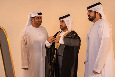 بورتريه لرجل عربي خليجي إماراتي يرتدي البشت الأسود المطرز بالذهبي بمساعدة صديقين، الاستعداد للمناسبات الرسمية والاحتفالات الوطنية، الاعتزاز بالهوية والتراث الإماراتي الأصيل ، مفهوم الأناقة والتراث والفخامه