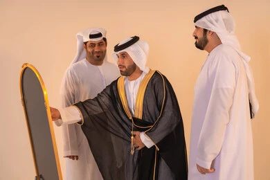الاستعداد للمناسبات الرسمية والاحتفالات الوطنية،الاعتزاز بالهوية والتراث الإماراتي الأصيل ، بورتريه لرجل عربي خليجي إماراتي يرتدي البشت الأسود المطرز بالذهبي بمساعدة صديقين،  مفهوم الأناقة والتراث والفخامه