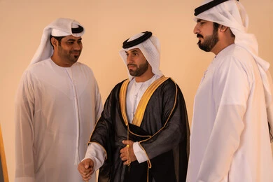 Celebrating national and heritage occasions, pride in the authentic Emirati identity and heritage, a portrait of an Arab Gulf Emirati man wearing a black bisht embroidered with gold with the help of two friends, the concept of elegance, heritage, and luxury.