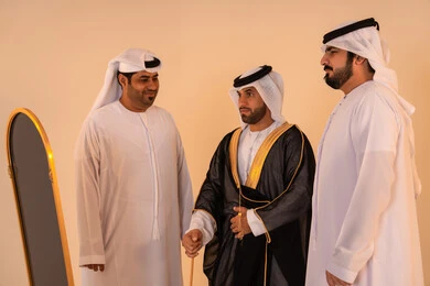 Aspects of authenticity and luxury in traditional Emirati attire, pride in the identity and authentic Emirati heritage, a portrait of an Arab Gulf Emirati man wearing a black bisht embroidered with gold with the help of two friends, preparing for official occasions and national celebrations.