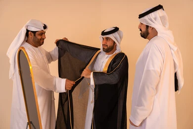Pride in the authentic Emirati identity and heritage, a portrait of an Arab Gulf Emirati man wearing a black bisht embroidered with gold with the help of two friends, preparing for official occasions and national celebrations, the concept of elegance, heritage, and luxury.