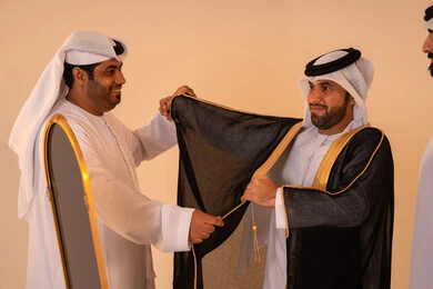 The concept of elegance, heritage, and luxury, pride in the identity and authentic Emirati heritage, a portrait of an Arab Gulf Emirati man wearing a black bisht embroidered with gold with the help of two friends, preparing for official occasions and national celebrations.