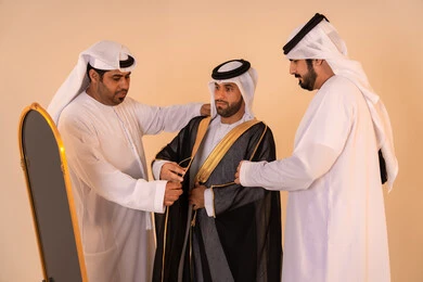 Aspects of authenticity and luxury in traditional Emirati attire, pride in the identity and authentic Emirati heritage, a portrait of an Arab Gulf Emirati man wearing a black bisht embroidered with gold with the help of two friends, preparing for official occasions and national celebrations.
