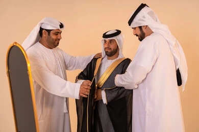 الاستعداد للمناسبات الرسمية والاحتفالات الوطنية،الاعتزاز بالهوية والتراث الإماراتي الأصيل ، بورتريه لرجل عربي خليجي إماراتي يرتدي البشت الأسود المطرز بالذهبي بمساعدة صديقين،  مفهوم الأناقة والتراث والفخامه