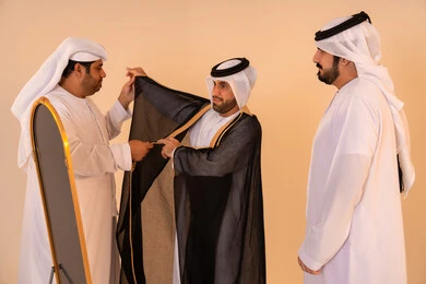 A portrait of an Emirati Gulf Arab man wearing a black bisht embroidered with gold, assisted by two friends, preparing for official occasions and national celebrations, expressing pride in the authentic Emirati identity and heritage, the concept of elegance, heritage, and luxury.
