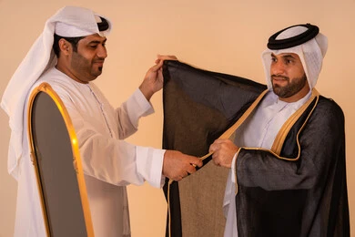 Aspects of authenticity and luxury in traditional Emirati attire, pride in the identity and authentic Emirati heritage, a portrait of an Arab Gulf Emirati man wearing a black bisht embroidered with gold with the help of his friend, preparing for official occasions and national celebrations.