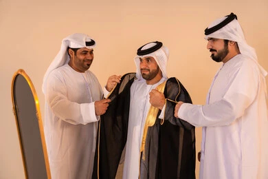 Pride in the authentic Emirati identity and heritage, a portrait of an Arab Gulf Emirati man wearing a black bisht embroidered with gold with the help of two friends, preparing for official occasions and national celebrations, the concept of elegance, heritage, and luxury.