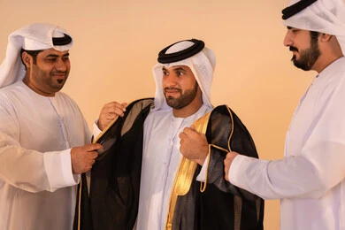 بورتريه لرجل عربي خليجي إماراتي يرتدي البشت الأسود المطرز بالذهبي بمساعدة صديقين، الاستعداد للمناسبات الرسمية والاحتفالات الوطنية، الاعتزاز بالهوية والتراث الإماراتي الأصيل ، مفهوم الأناقة والتراث والفخامه