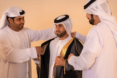 The concept of elegance, heritage, and luxury, pride in the identity and authentic Emirati heritage, a portrait of an Arab Gulf Emirati man wearing a black bisht embroidered with gold with the help of two friends, preparing for official occasions and national celebrations.