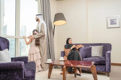 ايماءات السعادة و السرور، مشاهدة المسلسلات والأفلام الممتعة، الاستمتاع في قضاء الوقت مع الأهل، امراه عربيه خليجيه اماراتية ترتدي العباءة و الحجاب تجلس فى غرفه المعيشة تمسك ريموت التحكم ، اب يقف بجانب النافذة يقوم بمشاهده المناظر الطبيعية برفقة ابنته