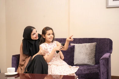 الاشارة الى شيء ما، ايماءات السعاده و السرور، أجواء اسرية سعيدة في المنزل، أم عربية خليجية اماراتية ترتدي العباءة والحجاب تجلس على الاريكه تقوم بمشاهدة التلفاز برفقه ابنتها، قضاء اوقات عائلية ممتعة، متابعة الافلام والمسلسلات