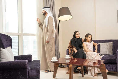 الاستمتاع بمشاهده المناظر الطبيعية، رجل عربي خليجي اماراتي يقف امام النافذة، مشاركة الوقت مع الابناء، ام عربيه خليجيه اماراتية تجلس على الاريكة برفقة ابنتها يقومون بمشاهدة التلفاز