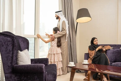 مشاهدة المسلسلات والأفلام الممتعة، الاستمتاع في قضاء الوقت مع الأهل، امراه عربيه خليجيه اماراتية ترتدي العباءة و الحجاب تجلس فى غرفه المعيشة تمسك ريموت التحكم ، اب يقف بجانب النافذة يقوم بمشاهده المناظر الطبيعية برفقة ابنته ، ايماءات السعادة و السرور