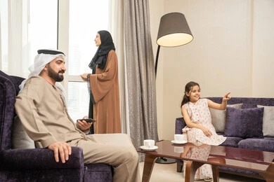 متابعة المسلسلات والأفلام المسلية، الاستمتاع في قضاء الوقت مع الأهل، رجل عربي خليجي اماراتي يرتدي الكندورة و الغتره يجلس فى غرفه المعيشة برفقة ابنته يمسك بيدية ريموت التحكم ، امراه عربيه خليجيه اماراتية تقف بجانب النافذة تقوم بمشاهده المناظر الطبيعية ، ايماءات السعادة و السرور