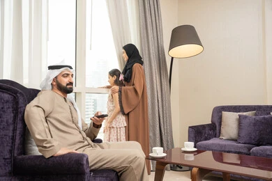 الاستمتاع في قضاء الوقت مع الأهل، رجل عربي خليجي اماراتي يرتدي الكندورة و الغتره يجلس فى غرفه المعيشة يمسك بيدية ريموت التحكم ، امراه عربيه خليجيه اماراتية تقف بجانب النافذة برفقة ابنتها ، ايماءات السعادة و السرور
