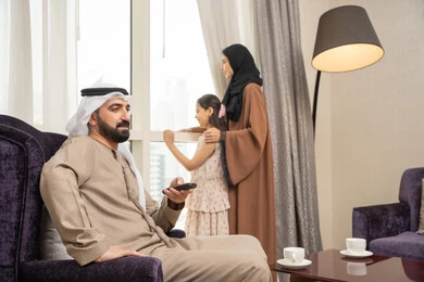 قضاء وقت عائلى ممتع، رجل عربي خليجي اماراتي يرتدي الكندورة و الغتره يجلس فى غرفه المعيشة يمسك بيدية ريموت التحكم ، امراه عربيه خليجيه اماراتية تقف بجانب النافذة برفقة ابنتها ، ايماءات السعادة و السرور