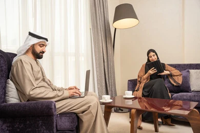Completing tasks from home, browsing social media, integrating technology into daily routines, an Arab Gulf Emirati man wearing a kandura and ghutrah sitting in the living room with his wife using a laptop and a tablet, expressions of focus and joy.