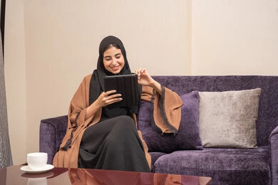 The use of modern devices and technologies, communicating with family and friends, sipping on a favorite drink, an Arab Gulf Emirati woman in a hijab wearing an abaya sitting on the couch enjoying moments of calm and relaxation, a Saudi woman making a video call via tablet with gestures of engagement.