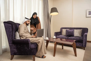 مشاركة الأفكار وإجراء النقاشات، ايماءات السعادة و السرور، الاشارة الي شاشة الحاسوب المحمول ، رجل عربي خليجي اماراتي يرتدي الكندورة و الغتره يجلس فى غرفه المعيشة برفقة زوجته يستخدم اللابتوب