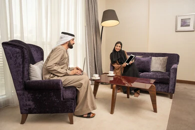 Gestures of happiness and joy, exchanging ideas and discussions, using modern technological devices, an Arab Gulf Emirati man wearing a kandura and ghutrah sitting in the living room with his wife using a laptop and a tablet.