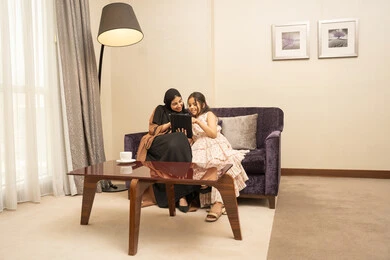 Monitoring online e-learning platforms, integrating learning with technology, watching recreational and educational activities, parents' interest in their children, an Arab Gulf Saudi woman wearing a hijab and abaya sitting on the couch with her daughter using a tablet.