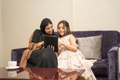 Watching recreational and educational activities, parents' interest in their children, following online educational platforms, integrating learning with technology, an Arab Gulf Saudi woman wearing a hijab and abaya sitting on the couch with her daughter using a tablet.