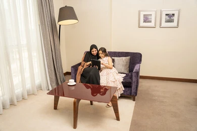 اهتمام الآباء بالأبناء، متابعة منصات التعليم الالكتروني عبر الانترنت، دمج التعلم بالتقنية والتكنولوجيا، مشاهدة الأنشطة الترفيهية والتعليمية، امراه عربية خليجية سعودية ترتدي الحجاب والعباءة تجلس على الاريكة برفقة ابنتها تستخدم جهاز التابلت