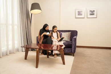 الاستمتاع بتناول المشروبات المفضلة، متابعة منصات التعليم الالكتروني عبر الانترنت، اهتمام الآباء بالأبناء، مشاهدة الأنشطة الترفيهية والتعليمية، امراه عربية خليجية سعودية ترتدي الحجاب والعباءة تجلس على الاريكة برفقة ابنتها تستخدم جهاز التابلت