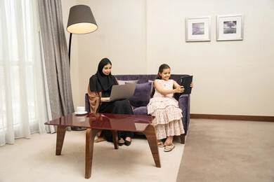 Making a video call via the tablet, a Gulf Arab Saudi woman wearing an abaya and hijab sitting on the sofa with her daughter using a laptop, the concept of remote work, spending quality time with children, gestures of happiness and joy.