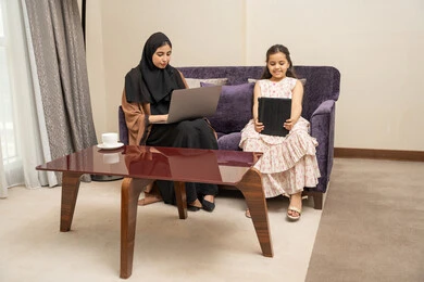 اجراء مكالمة فيديو عبر التابلت ، امراه عربيه خليجيه سعودية ترتدي العباءة و الحجاب تجلس على الاريكة برفقة ابنتها تقوم باستخدام الحاسب المحمول، مفهوم العمل عن بعد ، قضاء وقت ممتع مع الابناء ، ايماءات السعادة و السرور