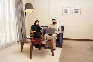 Completing daily tasks, gestures of focus and joy, the concept of remote work, integrating technology and tech into the daily routine, having a good time, an Emirati Arab couple in traditional attire sitting on the couch working on a laptop and a tablet.
