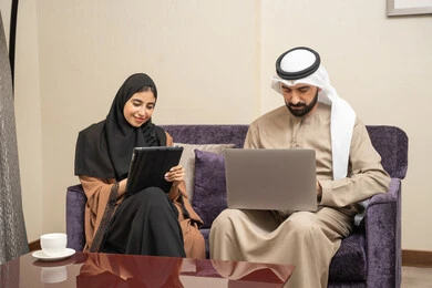ايماءات التركيز والسرور، مفهوم العمل عن بعد، دمج التقنية والتكنولوجيا في الروتين اليومي، قضاء وقت ممتع ، إنجاز المهام اليوميه، زوجان عربيان خليجيان اماراتيان يرتديان الزي التقليدي يجلسان على الاريكة يعملان على الحاسب المحمول وجهاز التابلت
