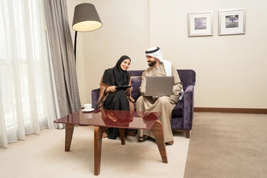 دمج التقنية والتكنولوجيا في الروتين اليومي، مفهوم العمل عن بعد، ايماءات التركيز والسرور، قضاء وقت ممتع ، إنجاز المهام اليوميه، زوجان عربيان خليجيان اماراتيان يرتديان الزي التقليدي يجلسان على الاريكة يعملان على الحاسب المحمول وجهاز التابلت