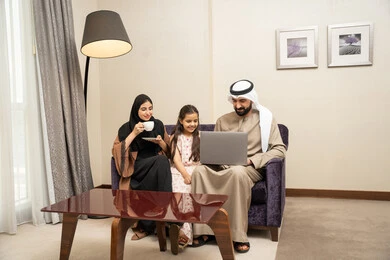 قضاء وقت ممتع مع الأبناء، مشاهدة الافلام والبرامج الترفيهيه، رجل عربي خليجي اماراتي يرتدي الكندورة و الغتره يجلس على الكنبة بجانب ابنته وزوجته يقومون باجراء مكالمة فيديو عبرالحاسب المحمول، الاستمتاع بتناول المشروبات المفضلة