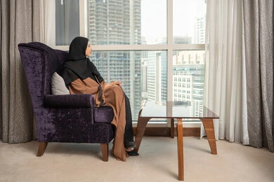 قضاء وقت ممتع، صورة من الجانب لامراة عربيه خليجيه سعودية ترتدي العباءة و الحجاب تجلس في غرفة المعيشة بجانب النافذة، الجلوس والاستمتاع بالمناظر الخارجيه