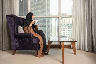 Enjoying hot drinks, having a good time, a side view of an Arab Gulf Saudi woman wearing an abaya and hijab sitting in the living room by the window holding a cup of coffee, drinking coffee and hot beverages.