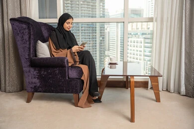 ايماءات السعادة و السرور، الدردشة مع الأهل والأصدقاء وتصفح مواقع التواصل الاجتماعي، استخدام الأجهزة التقنية الحديثة، صورة من الجانب لامراة عربيه خليجيه سعودية ترتدي العباءة و الحجاب تجلس في غرفة المعيشة بجانب النافذة تمسك الهاتف المحمول
