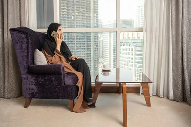 Making a voice call via mobile phone, using modern technological devices, a side view of a Saudi Gulf Arab woman sitting in the living room by the window holding a mobile phone, chatting and conversing with friends and relatives, gestures of happiness and joy.