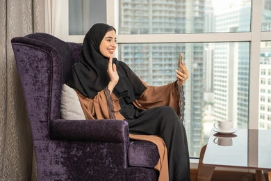 Documenting happy moments, taking a selfie with a mobile phone, a side view of a Saudi Gulf Arab woman sitting in the living room by the window holding her phone, raising her hand with gestures of peace and greeting, making a video call, the concept of live broadcasting.