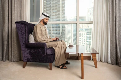 إدارة شؤون الأعمال وتنظيمها عبر اللابتوب، إنجاز مهام العمل عن بعد، استخدام جهاز تقني حديث ومتطور، صورة من الجانب لرجل عربي اماراتي خليجي يرتدي الكندورة والغترة الاماراتية يجلس في غرفة المعيشة بجانب النافذة يستخدم الحاسوب المحمول
