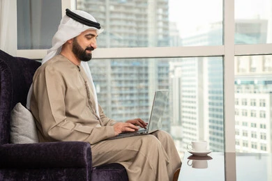 استخدام جهاز تقني حديث ومتطور، صورة من الجانب لرجل عربي اماراتي خليجي يرتدي الكندورة والغترة الاماراتية يجلس في غرفة المعيشة بجانب النافذة يستخدم الحاسوب المحمول، إنجاز مهام العمل عن بعد، إدارة شؤون الأعمال وتنظيمها عبر اللابتوب